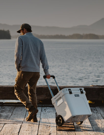 Man pulling a YETI cooler along a dock with a scenic lake and mountains in the background
