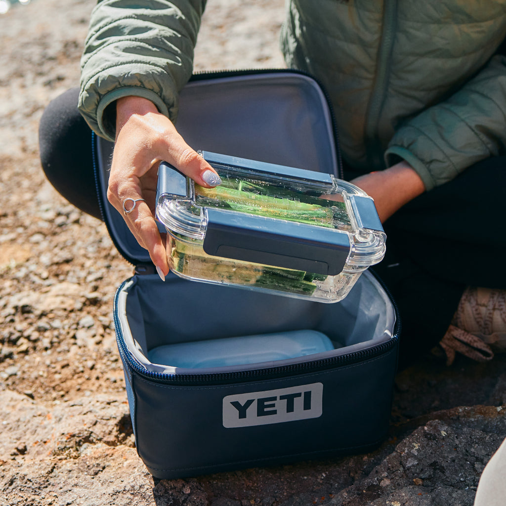 Person opening a YETI lunch box with a clear container inside on a rocky ground.