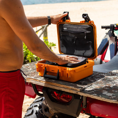 An orange YETI hard case with black mesh interior panels is open on the back of a red ATV at a beach, with a shirtless person placing a black item inside the case.