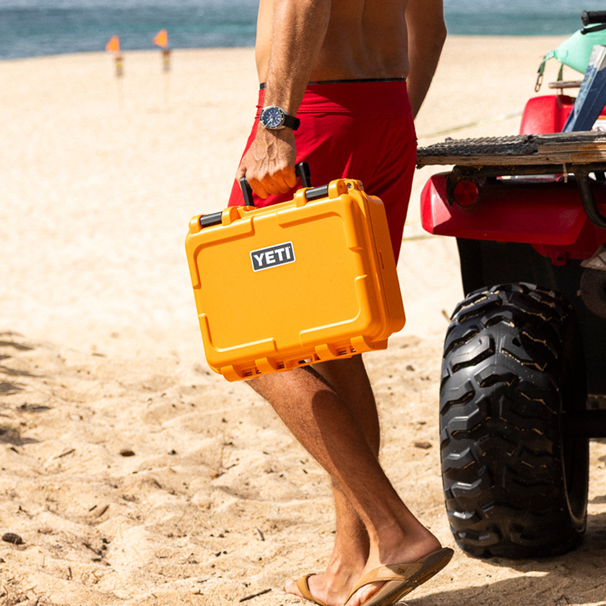 A man in red swim trunks carrying a bright yellow YETI hard case on a sandy beach next to an ATV.
