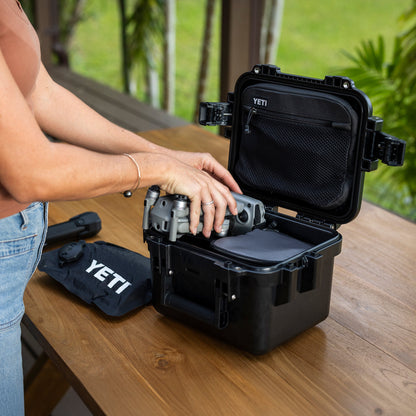 A person places a gray drone into an open black YETI hard case on a wooden table, with a black YETI bag nearby and a blurred outdoor background of greenery.