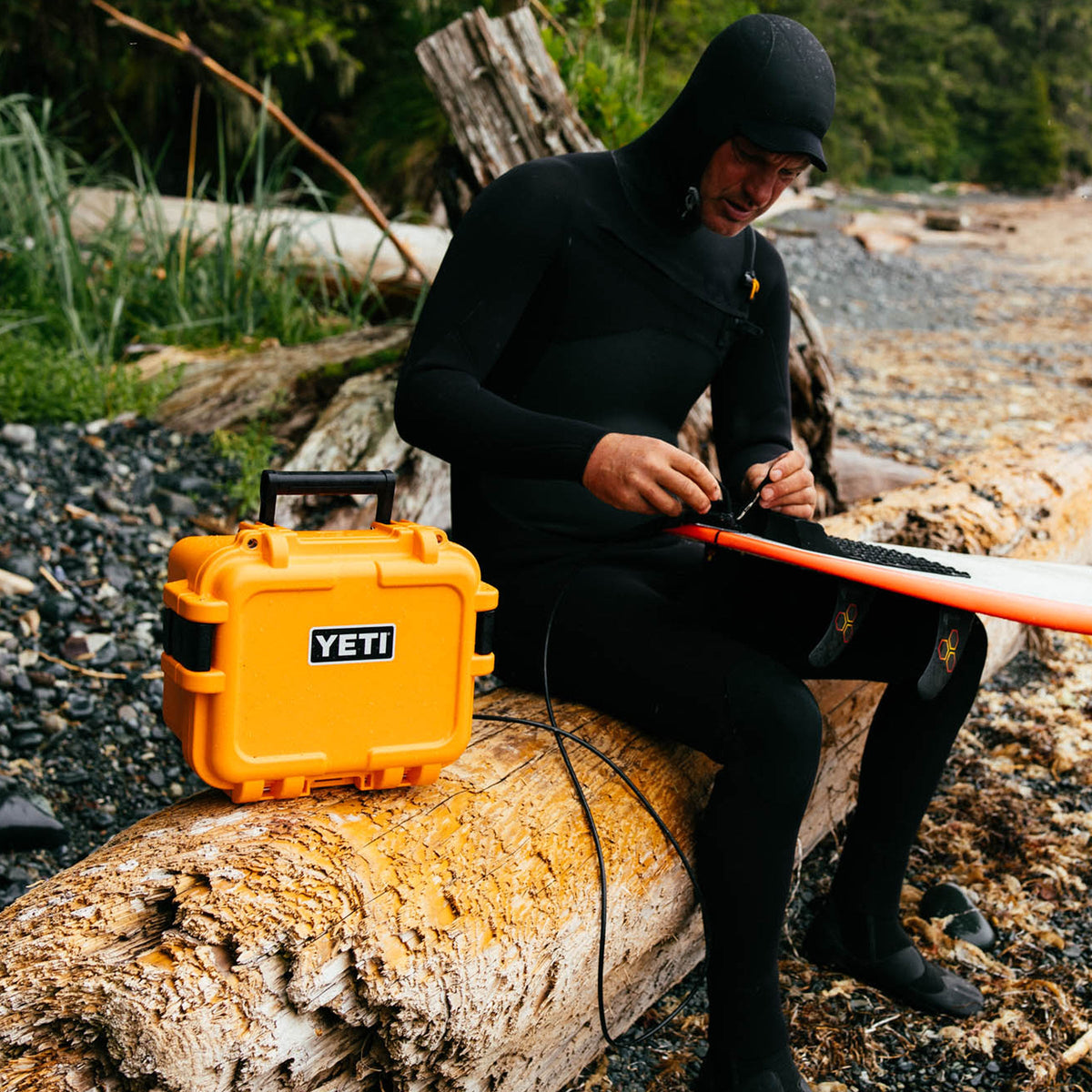 A person in a black wetsuit sits on a large log outdoors by the water, working on an orange surfboard. A bright yellow YETI hard case is placed beside them on the log.