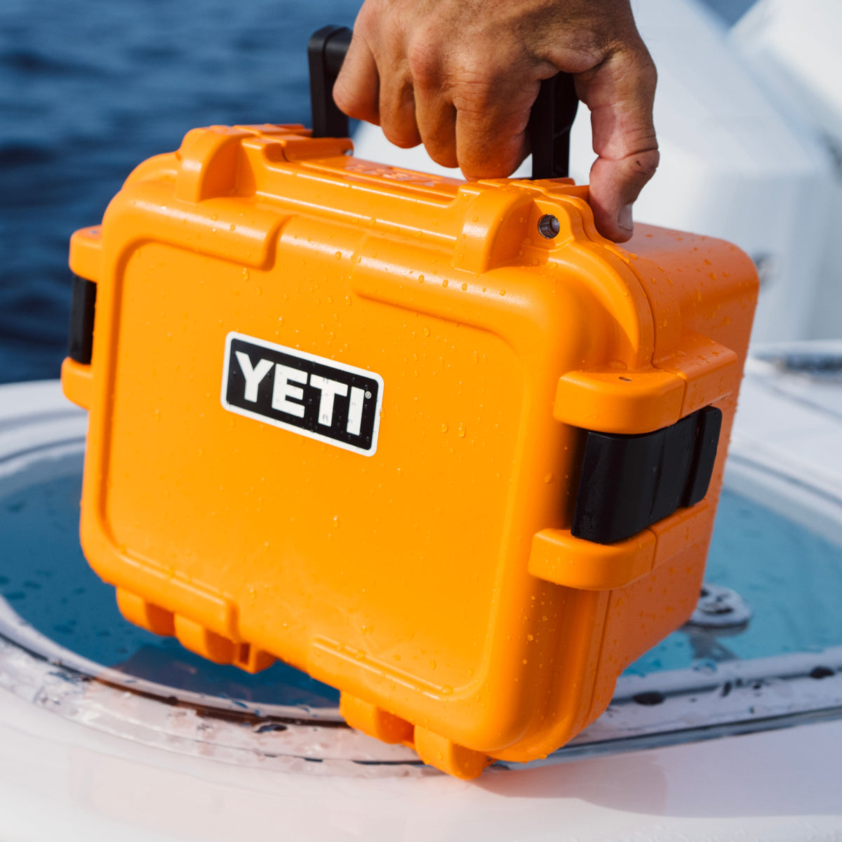 A bright orange YETI waterproof hard case with black latches and a black handle, being held by a person over the edge of a boat with water visible in the background.