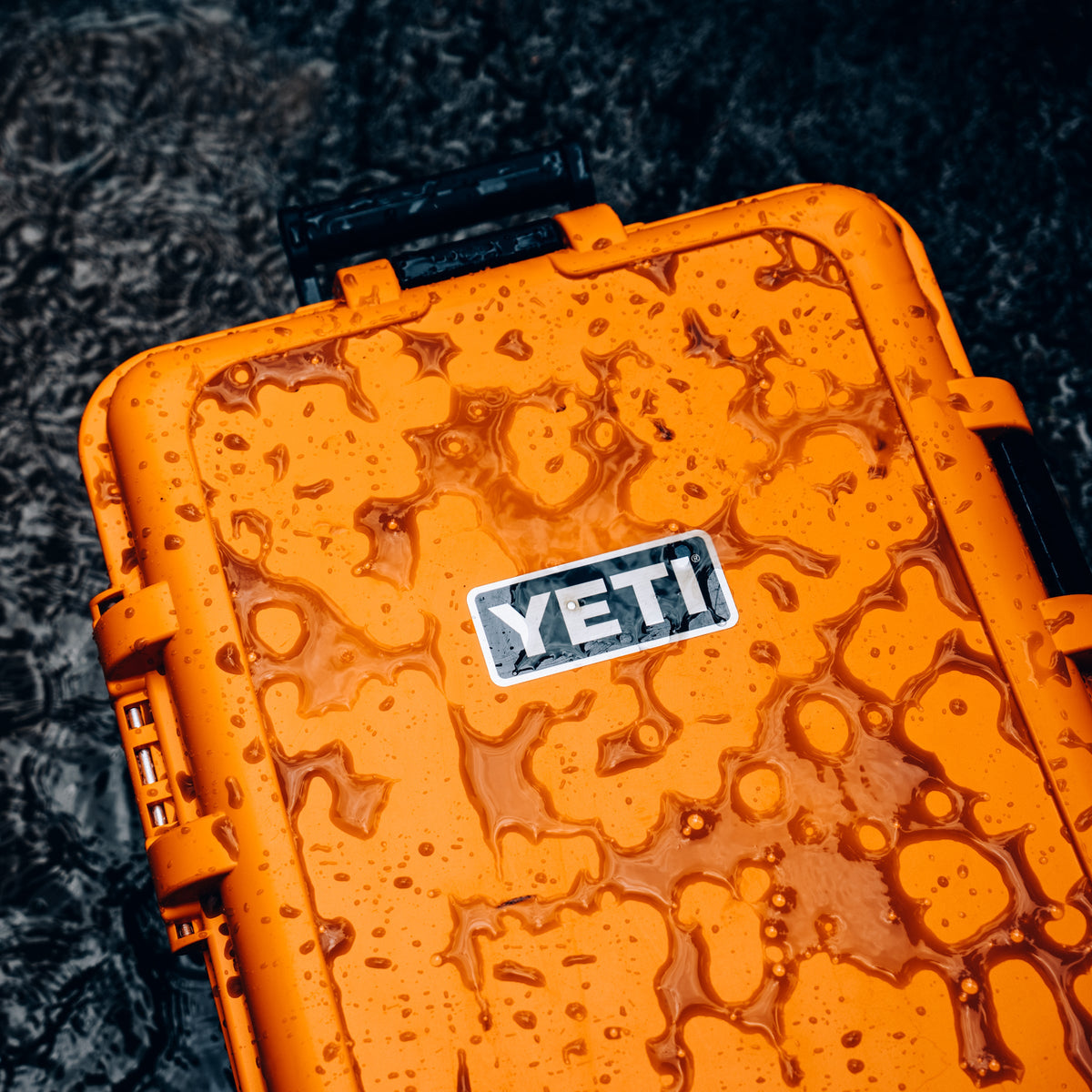 An orange YETI waterproof hard case covered in water droplets, with the YETI logo visible on the front, set against a dark textured background.