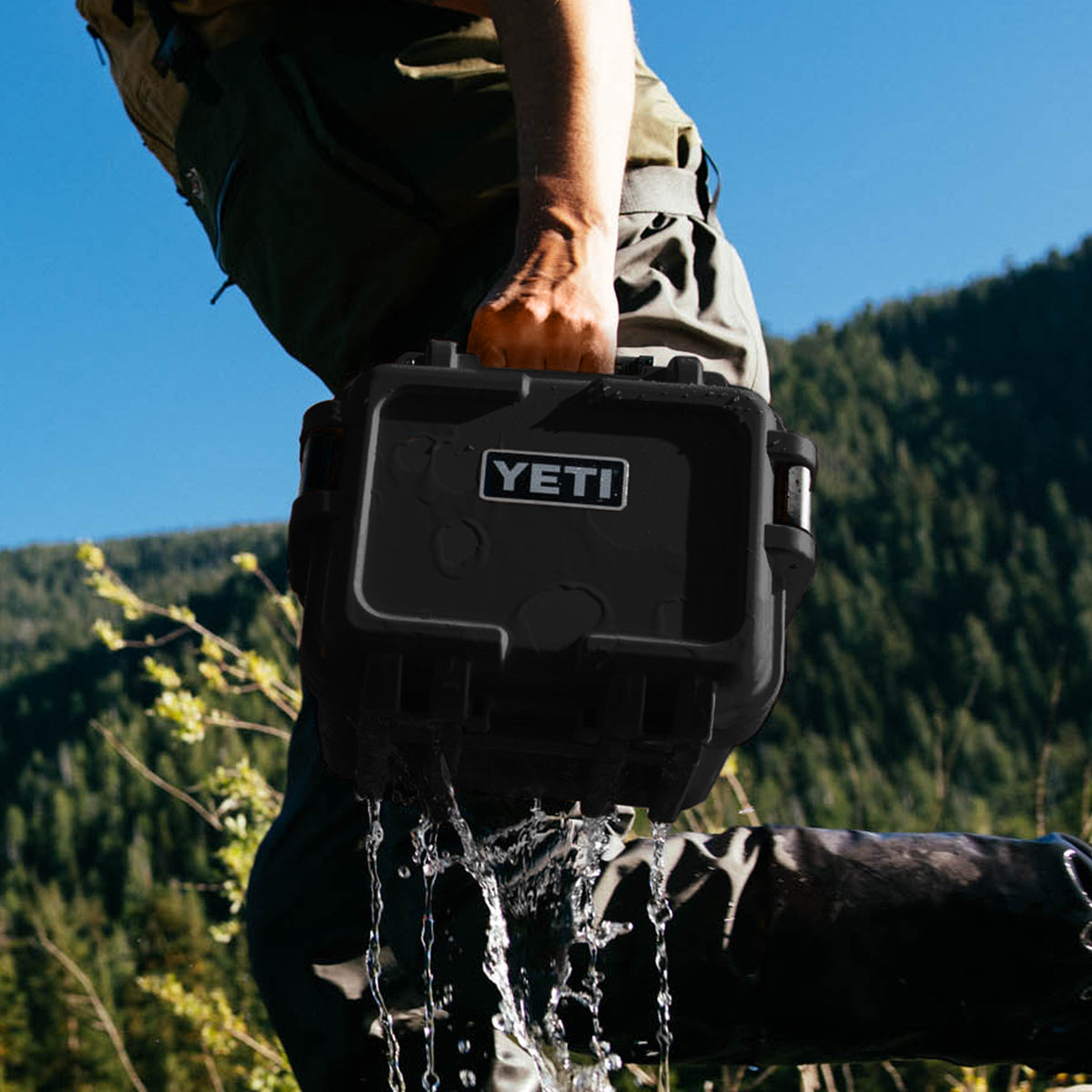 A person outdoors carrying a black YETI waterproof hard case while water pours from the bottom, with a forested mountain and blue sky in the background.