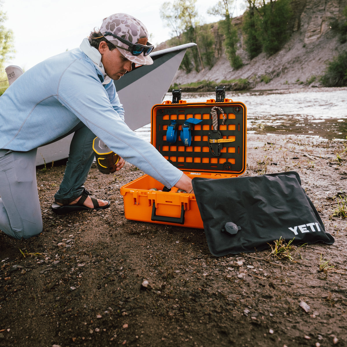 A man kneels on a muddy riverbank next to an open bright orange YETI hard case filled with gear, with a black YETI bag beside it and a boat visible in the background.