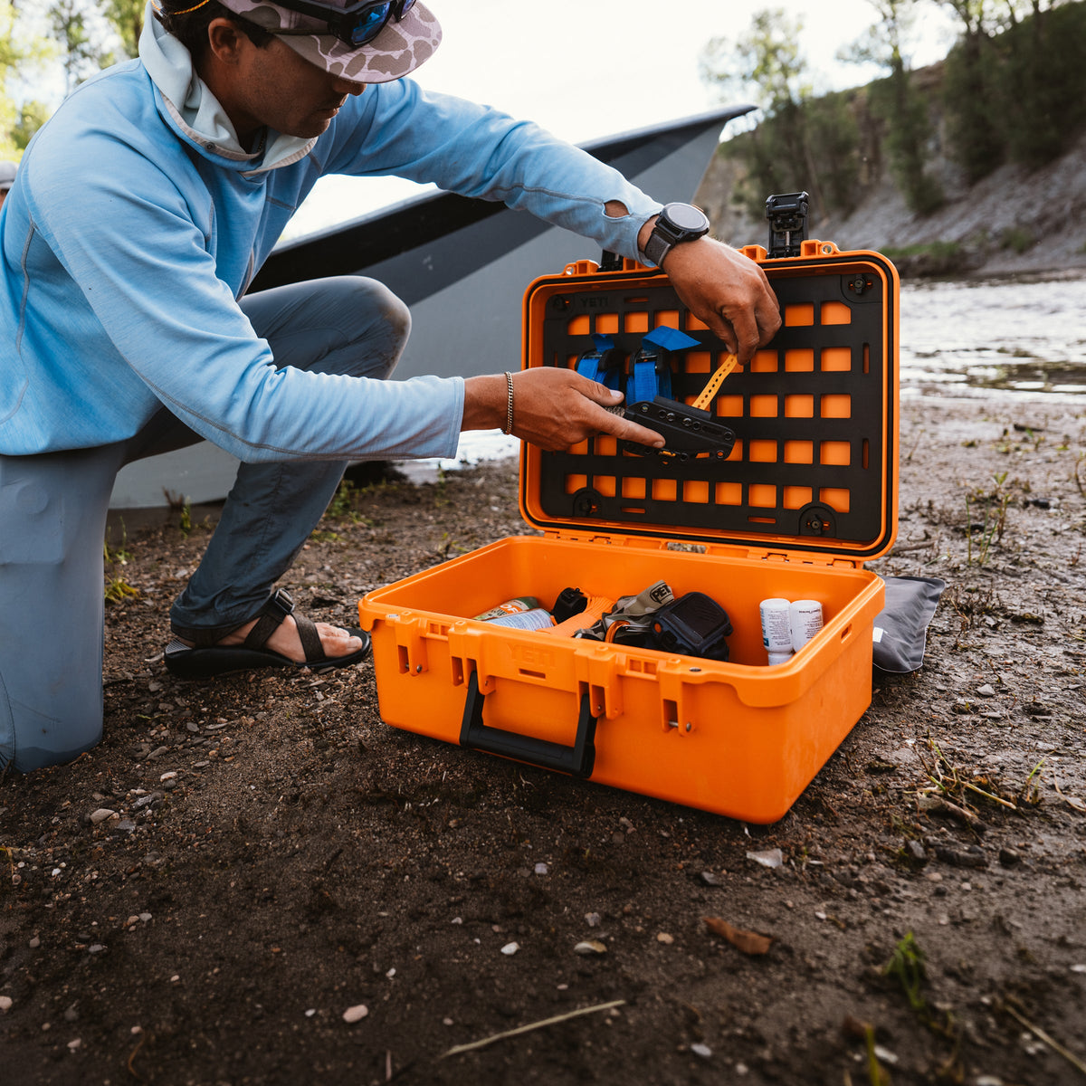 A man in a light blue long-sleeve shirt and jeans kneels beside an open bright orange hard case on a dirt shore, placing or adjusting a black and blue device inside the case.