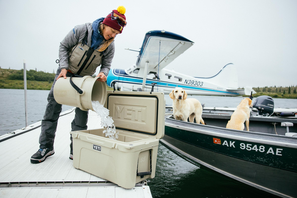 Person on a boat pouring ice from a bucket into a Yeti cooler with two dogs nearby.