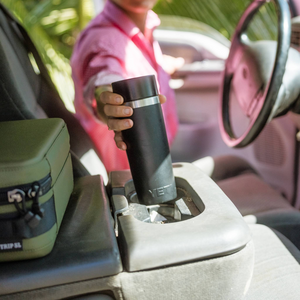 Person holding a black insulated mug inside a vehicle with a green box on the passenger seat.