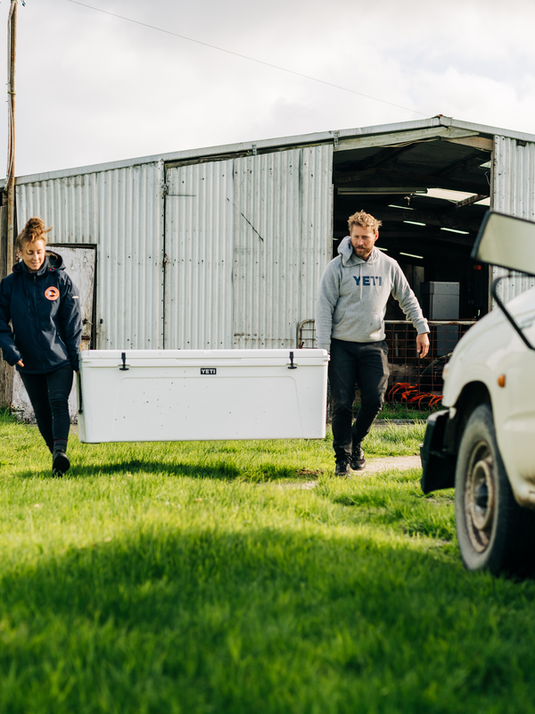 Two people walking with a large white YETI cooler outdoors near a building.